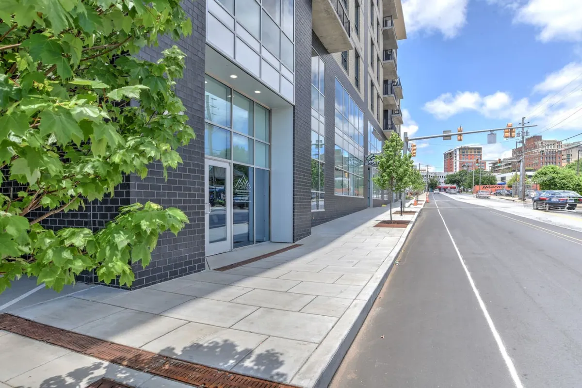This image shows a modern downtown streetscape with large glass windows, bold architectural lines, and lush green trees lining the sidewalk. Pedestrians can be seen enjoying a walk on a sunny day, and the vibrant setting highlights the accessibility and energy of the urban area.