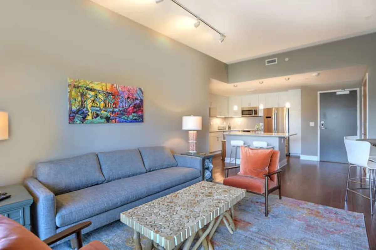 A spacious and colorful living area featuring vibrant wall art, a comfortable sofa, a coffee table, and a stylish lighting fixture. The scene suggests a welcoming space ideal for both relaxation and group activities, with a nod to artistic influences in its decor.