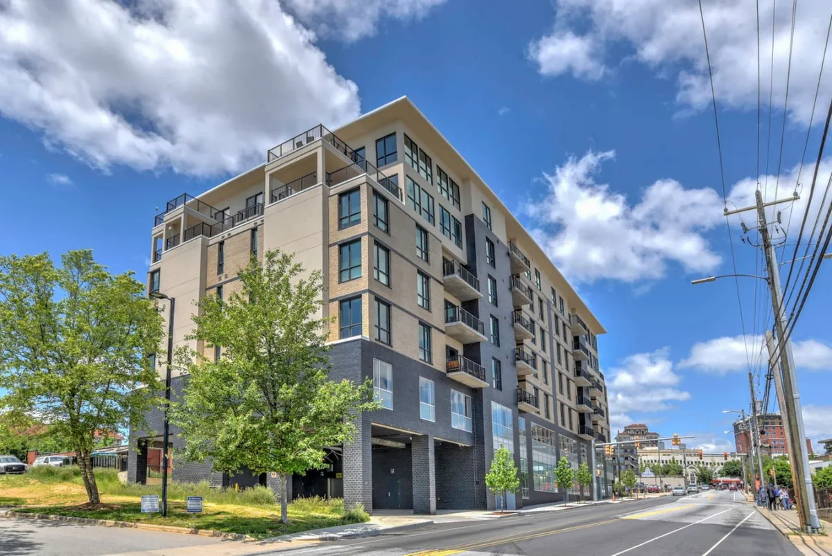 A contemporary exterior view of a modern building featuring large windows, balconies, and lush landscaping. The setting suggests an inviting urban environment, ideal for walking and socializing, with clear indications of proximity to nightlife and community events.