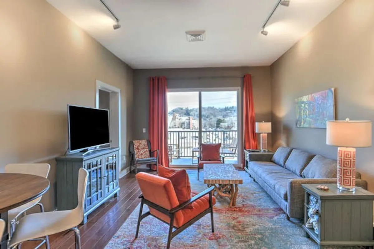 A welcoming open-concept space featuring a plush sofa, striking wall art, and a dining table near large windows. The sunlight fills the room, accentuating the cozy seating and offering a glimpse of the private balcony beyond. The layout encourages conversation and group gatherings.