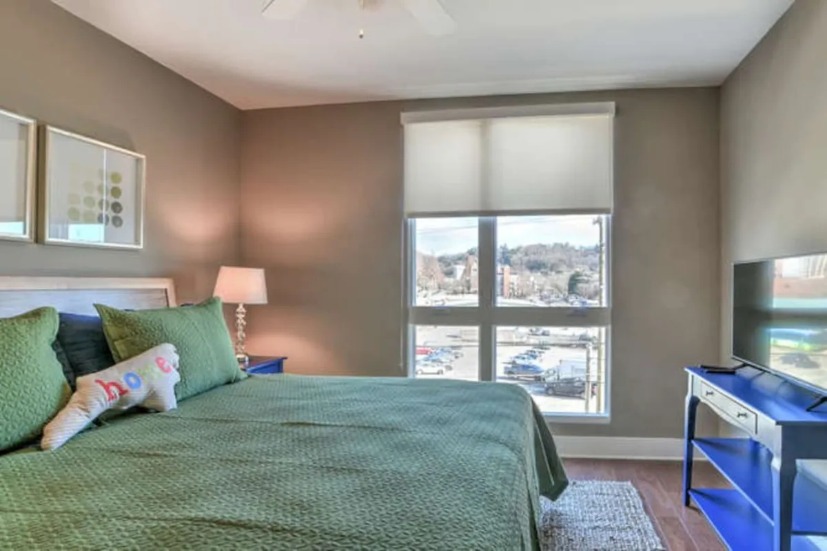 This image captures a bright, inviting bedroom space with green bedding, a window offering a view outside, and blue furniture. The placement of the window and the sense of light suggest a connection to the outdoors, evoking the idea of starting an exciting day in Asheville or relaxing after exploring the city’s nightlife.