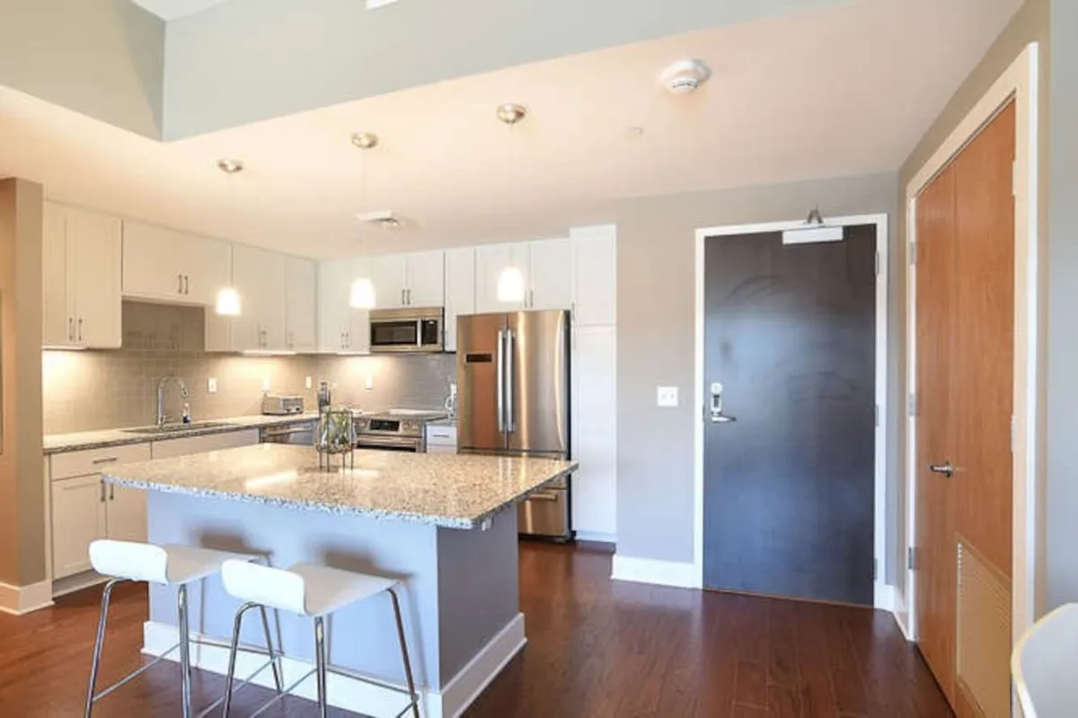 The image features a sleek, modern elevator lobby with island countertop and stainless steel accents, illuminated by pendant lighting. Its clean lines and inviting atmosphere suggest seamless movement through the building’s common areas.