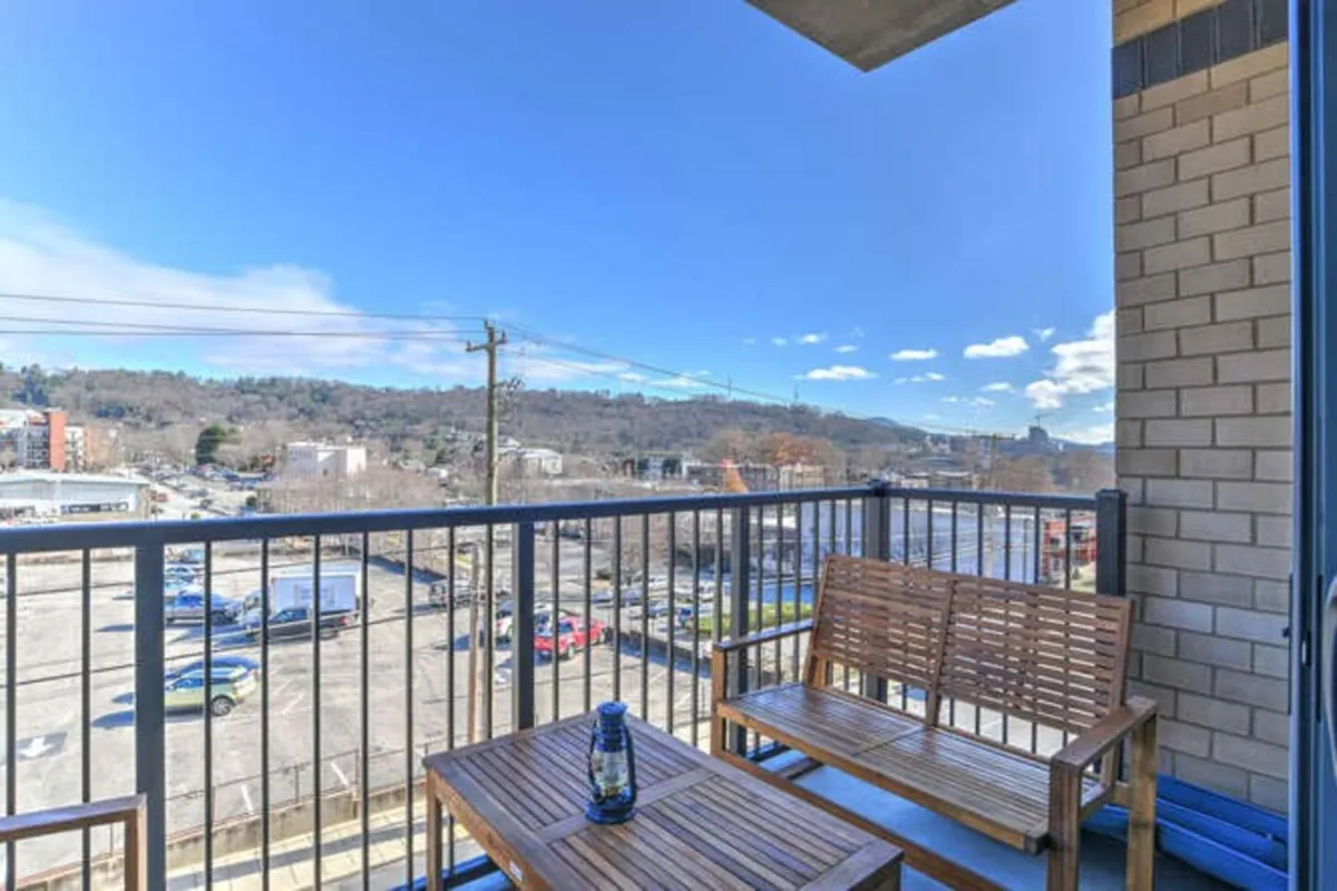 A tranquil private balcony with wooden bench seating, offering views of the city skyline and blue sky. The space is ideal for morning coffee or unwinding at sunset, seamlessly extending the living area outdoors.